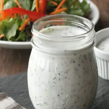 Mason jar of creamy, herb-speckled homemade ranch dressing on a dark slate board, with a blurred background of fresh green salad.