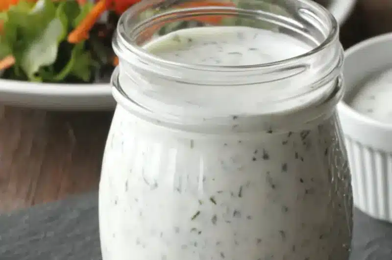 Mason jar of creamy, herb-speckled homemade ranch dressing on a dark slate board, with a blurred background of fresh green salad.