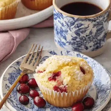 Single powdered cranberry orange muffin on blue transferware plates next to a coffee mug, highlighting the vibrant cranberries.