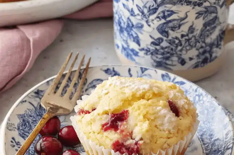 Single powdered cranberry orange muffin on blue transferware plates next to a coffee mug, highlighting the vibrant cranberries.