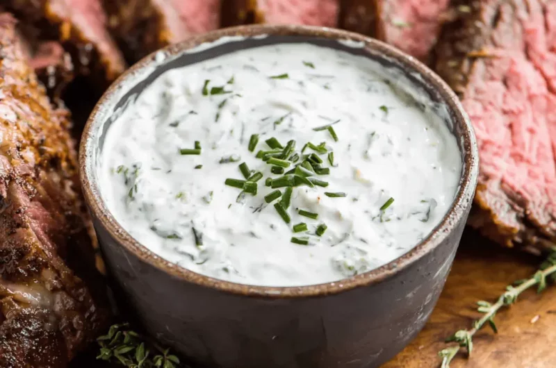 Close-up of a rich Horseradish Cream Sauce in a rustic bowl, garnished with chives, served alongside medium-rare roasted beef.