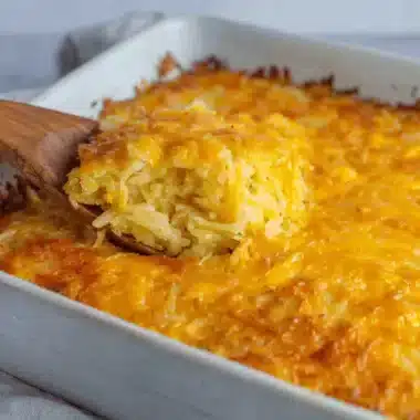 Ultimate Foolproof Cheesy Hashbrown Casserole 4 A wooden spoon scoops a warm, golden Cheesy Hashbrown Casserole with bubbling melted cheese from a white baking dish.