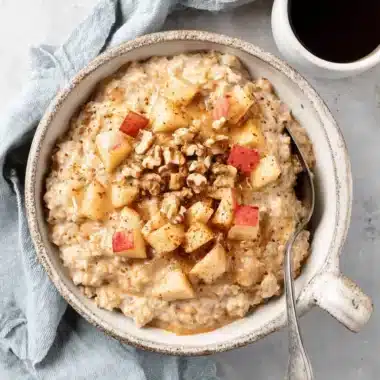 The Ultimate Apple Cinnamon Oatmeal Recipe 3 A rustic bowl of warm apple cinnamon oatmeal, topped with diced red apples, walnuts, and a sweet drizzle, served on a light surface.