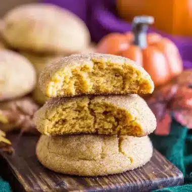 The Ultimate Chewy Pumpkin Snickerdoodles 3 Stack of warm, golden-brown Pumpkin Snickerdoodles, one broken to show its soft, orange interior, on a rustic wooden board with autumn leaves.