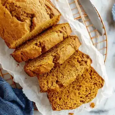 Freshly baked pumpkin bread loaf, partially sliced on a cooling rack with a knife.