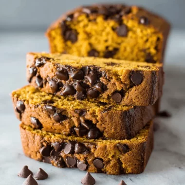 Stack of three moist slices of homemade pumpkin chocolate chip bread on a light surface, with the loaf in the background.