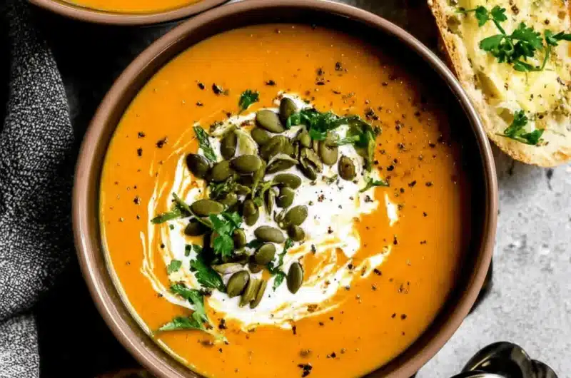 Warm pumpkin soup in two bowls, garnished with cream, pumpkin seeds, and fresh herbs, served with toasted garlic bread on a gray surface.