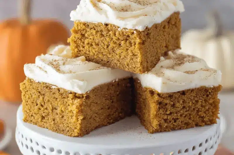 Three frosted pumpkin bars dusted with spice on a decorative white cake stand, surrounded by autumn pumpkins.
