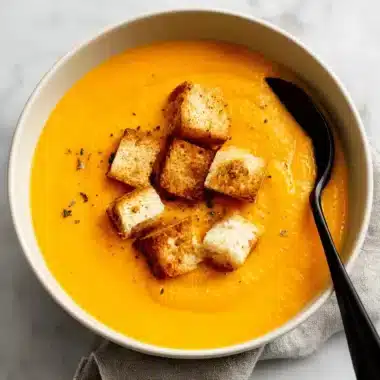 Overhead view of creamy sweet potato soup in a white bowl, garnished with crispy croutons and fresh herbs on a marble surface.