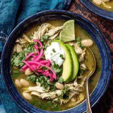 Hearty bowl of green chili chicken soup garnished with avocado, pickled onions, and fresh cilantro on a dark background.