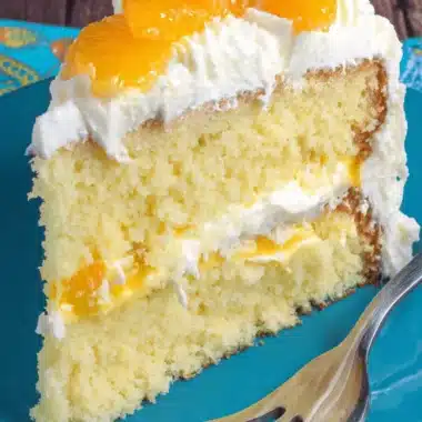 Amazing Mandarin Orange Cake: Quick & Moist 3 Delectable slice of layered mandarin orange cake with creamy filling, fresh segments, and whipped cream on a teal plate.