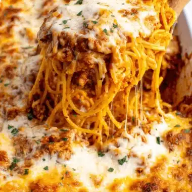 Ultimate Cheesy Baked Spaghetti Recipe 4 Overhead close-up of a wooden spoon scooping a portion of rich, gooey Cheesy Baked Spaghetti casserole from a white dish.