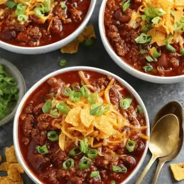 The Ultimate Foolproof Beef Chili Recipe 4 Hearty bowl of beef chili recipe topped with melted cheese, tortilla chips, and green onions on a dark background.