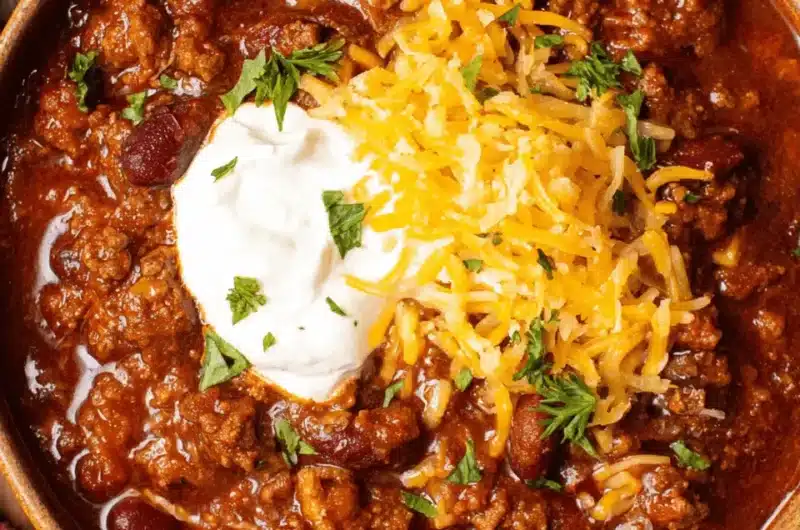 High-angle close-up of a rustic bowl filled with chunky, beef-laden texas chili, generously topped with shredded cheddar cheese, sour cream, and fresh cilantro.