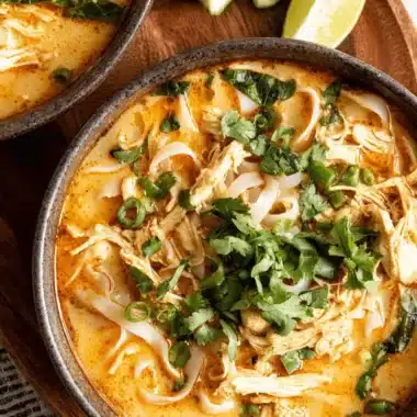 Rich, creamy Thai Chicken Soup with shredded chicken, wide rice noodles, cilantro, and lime wedges on a rustic wooden board.