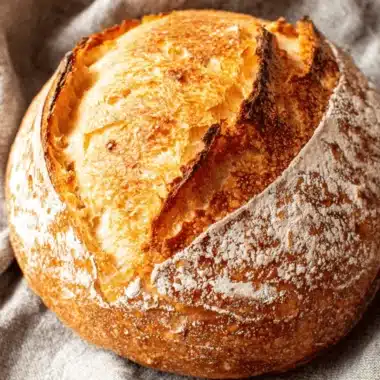 Amazing Overnight Sourdough Bread for Beginners 3 Rustic close-up of a perfectly baked boule of Overnight Sourdough Bread with a dark, caramelized crust and prominent ear, resting on a textured linen cloth.
