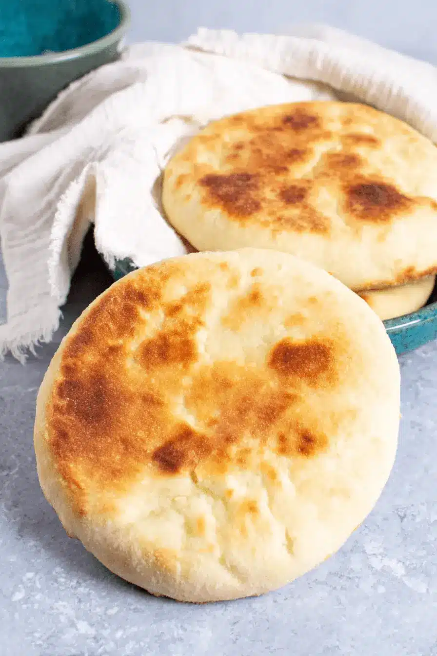 Foolproof Sourdough Pita Bread: Perfect Puff! 2 Freshly baked Sourdough Pita Bread with visible golden char marks, resting next to a stack in a vibrant teal ceramic bowl.