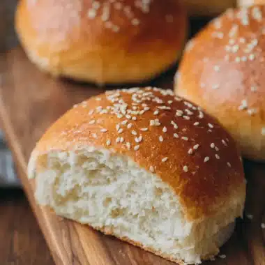 Freshly baked Sourdough Hamburger Buns topped with sesame seeds displayed on a rustic wooden board, showing the soft, airy crumb structure.
