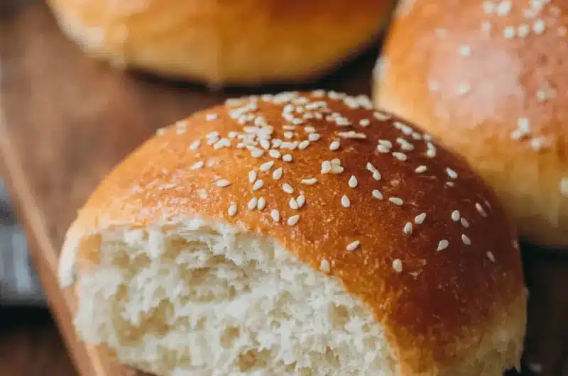 Freshly baked Sourdough Hamburger Buns topped with sesame seeds displayed on a rustic wooden board, showing the soft, airy crumb structure.
