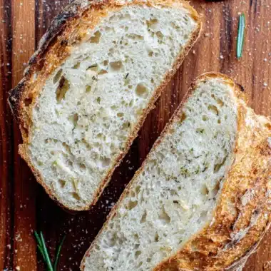 Ultimate Foolproof Parmesan Sourdough Bread 4 Top-down view of two slices of herb-infused Parmesan Sourdough Bread resting on a rustic wooden surface with fresh rosemary sprigs.