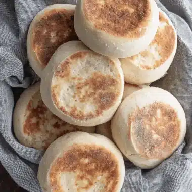 Simple Sourdough Discard English Muffins 3 A close-up, top-down view of freshly baked Sourdough Discard English Muffins piled on a gray cloth, highlighting the golden-brown tops and airy texture.