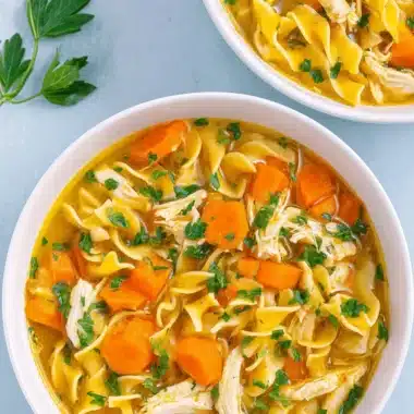 The Ultimate Comforting Chicken Noodle Soup Recipe 4 Overhead view of rich, golden Chicken Noodle Soup with wide egg noodles, shredded chicken, and carrots in white bowls.