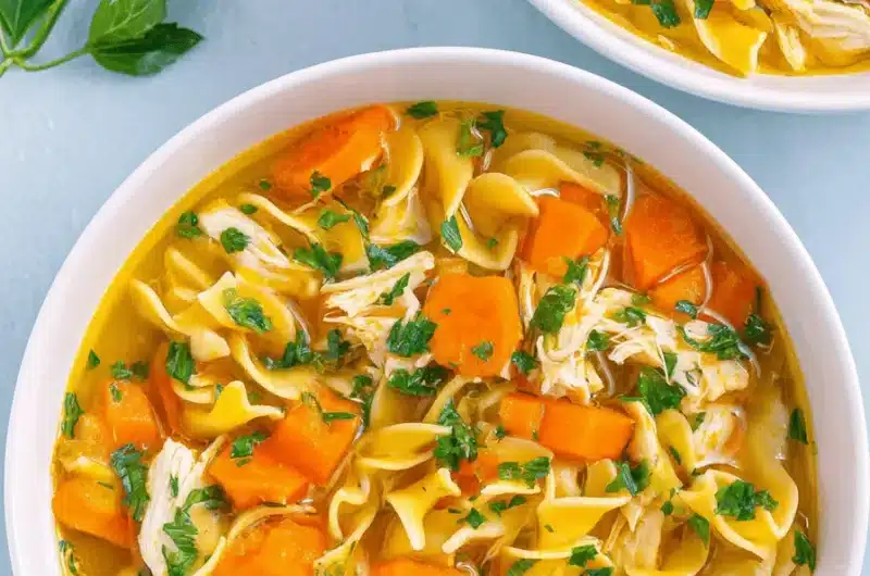 Overhead view of rich, golden Chicken Noodle Soup with wide egg noodles, shredded chicken, and carrots in white bowls.