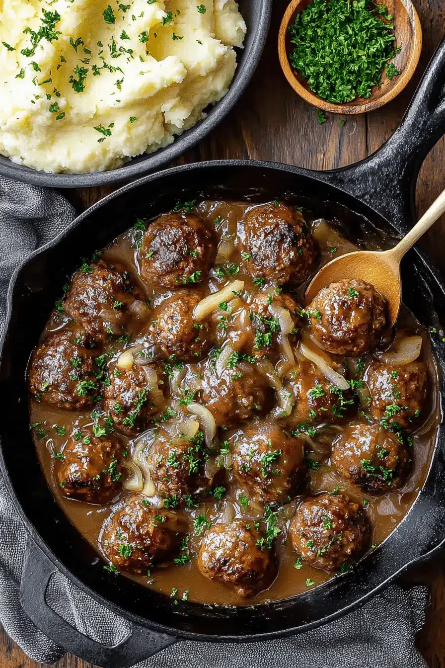 Amazing Salisbury Steak Meatballs in 40 Minutes 2 Cast-iron skillet with rich brown gravy, plump salisbury steak meatballs, and mashed potatoes garnished with parsley.
