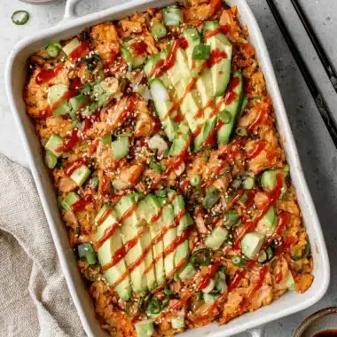 The Ultimate Salmon Sushi Bake Recipe 4 Vibrant, overhead shot of a prepared salmon sushi bake recipe in a white casserole dish, topped with fanned avocado and sriracha.