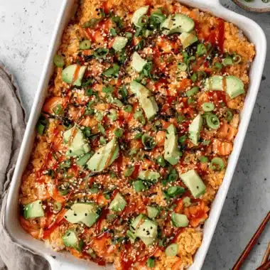 The Ultimate Salmon Sushi Bake Recipe 4 Top-down view of a golden-brown salmon sushi bake recipe with avocado, spicy mayo, and sesame seeds in a white dish.