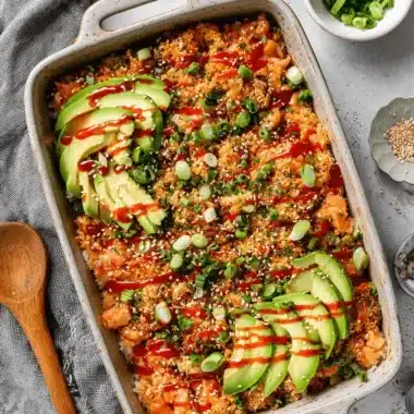 Amazing Salmon Sushi Bake Recipe for Busy Nights 3 A rectangular ceramic dish filled with a finished salmon sushi bake recipe, topped with fanned avocado, sriracha, and green onions.