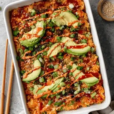 Ultimate Salmon Sushi Bake Recipe 3 Close-up of a golden-brown salmon sushi bake recipe in a white dish, topped with sliced avocado, creamy spicy sauce, green onions, and sesame seeds.