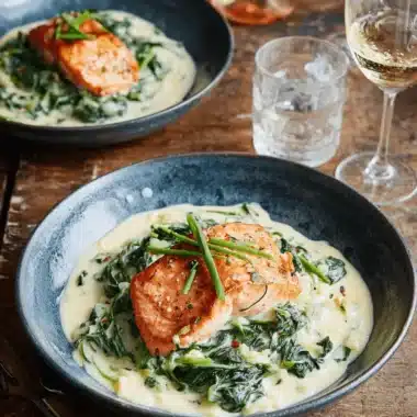 Amazing Baked Salmon with Spinach Recipe 3 Two servings of baked salmon with spinach and creamy sauce in dark bowls on a rustic wooden table, garnished with fresh chives.