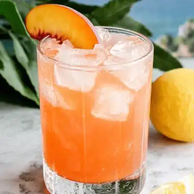 The Ultimate Peach Lemonade for Summer 3 Tall glass of vibrant Peach Lemonade with ice and a peach slice garnish, set on marble with lemons and a tropical seascape background.