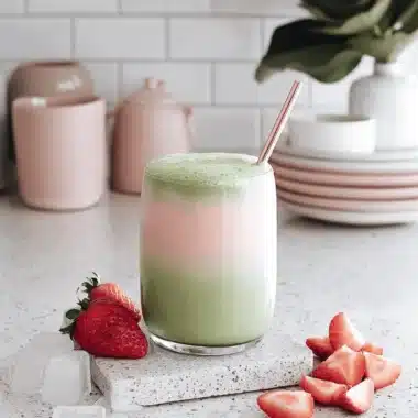 Easy & Beautiful Strawberry Matcha Milk 4 Layered Strawberry matcha Milk served in a clear glass with fresh strawberries and a reusable rose-gold straw on a bright kitchen counter.