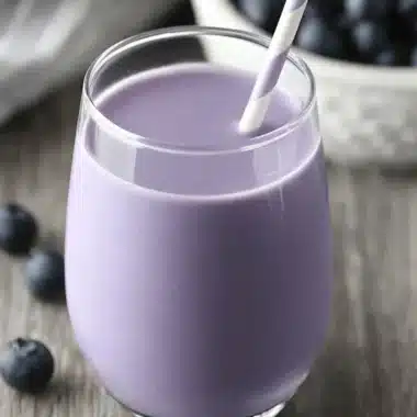 Vibrant Homemade Blueberry Milk Recipe 3 Vibrant purple blueberry milk in a clear glass with a striped straw, surrounded by fresh blueberries on a rustic wooden table.