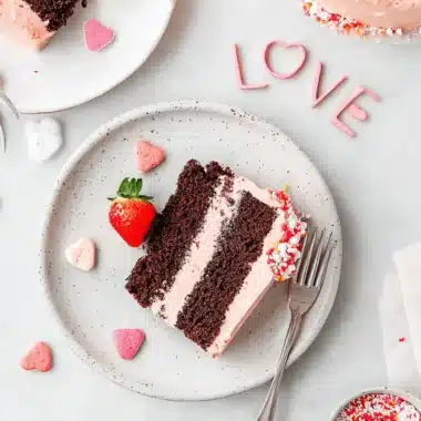 Stunning Valentine's Day Cake Recipe 3 Top-down image of a slice of layered chocolate valentine's day cake featuring pink frosting, heart candies, and fresh strawberries on a bright surface.