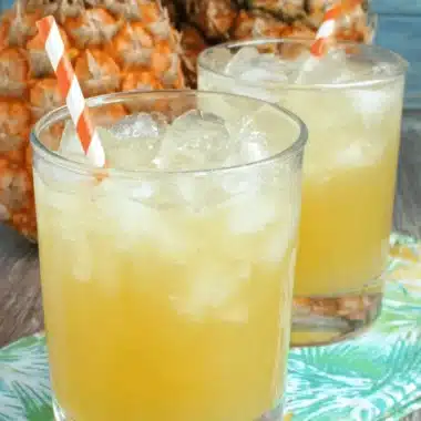 Easy Pineapple Lemonade Punch for Any Crowd 3 Two glasses of frothy pineapple lemonade punch served over ice with festive striped straws next to fresh pineapples.