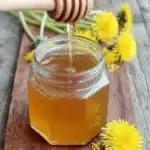 Magic Dandelion Honey Recipe: Vegan Sweetness 8 Hexagonal glass jar of golden honey on a wooden board with scattered dandelions, illustrating the final product of the dandelion honey recipe.