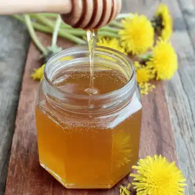 Magic Dandelion Honey Recipe: Vegan Sweetness 3 Hexagonal glass jar of golden honey on a wooden board with scattered dandelions, illustrating the final product of the dandelion honey recipe.