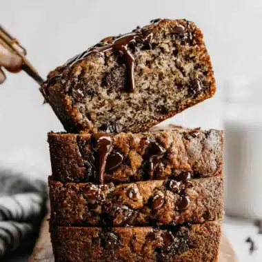 Ultimate Moist Chocolate Chip Banana Bread 4 Three moist slices of Chocolate Chip Banana Bread stacked on a wooden board, drizzled with chocolate sauce, with one slice being lifted.