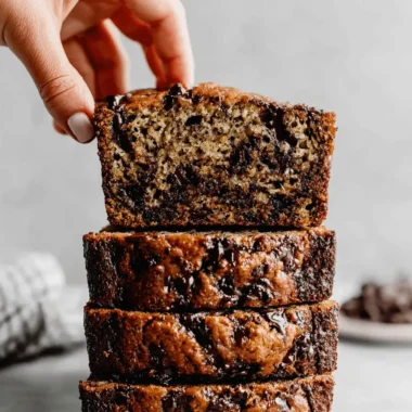 Unbelievably Moist Chocolat Banana Bread 3 Hand reaching for a slice of moist, stacked chocolat banana bread with dark chocolate chips.