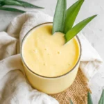 Easy & Vibrant Mango Pineapple Smoothie 8 Overhead close-up of a creamy, vibrant yellow mango pineapple smoothie in a clear glass, garnished with fresh pineapple leaves.