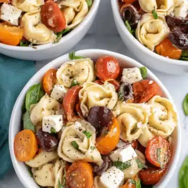 Quick & Easy Cheese Tortellini Pasta Salad 3 Overhead view of three bowls of vibrant cheese tortellini pasta salad with cherry tomatoes, Kalamata olives, feta, and fresh basil on a light surface.