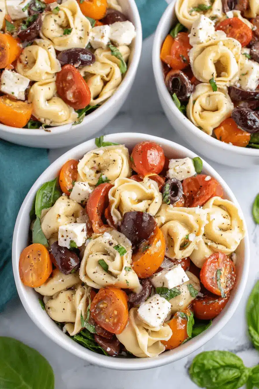 Quick & Easy Cheese Tortellini Pasta Salad 2 Overhead view of three bowls of vibrant cheese tortellini pasta salad with cherry tomatoes, Kalamata olives, feta, and fresh basil on a light surface.