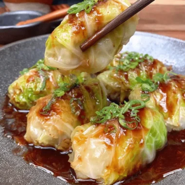 Amazing Homemade Cabbage Dumplings Recipe 3 Close-up of vibrant green cabbage dumplings in a dark ceramic bowl, glistening with savory sauce and topped with green onions, held by chopsticks.