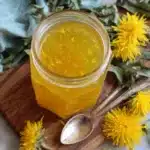 Unlock the Magic of Fresh Dandelion Jelly 7 Jar of shimmering golden Fresh Dandelion Jelly on a rustic wooden board, framed by fresh yellow dandelion blooms and green leaves.