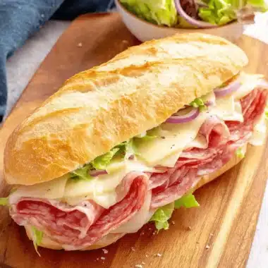 Ultimate Baked Italian Grinder Sandwiches 3 Freshly prepared italian grinder sandwiches on a rustic wooden board, filled with salami, provolone, lettuce, and red onion.