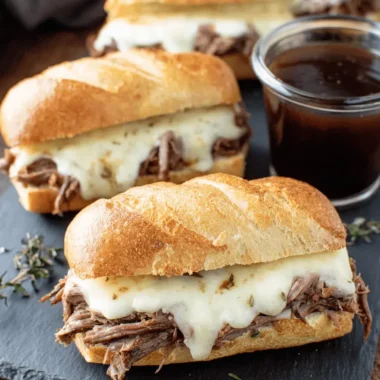 Amazing Crockpot French Dip Sandwiches 4 Close-up of homemade crockpot french dip sandwiches with provolone cheese and a side of au jus on a dark slate board.