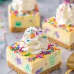Ultimate Funfetti Cheesecake Bars: Easy Recipe 8 Three colorful Funfetti Cheesecake Bars, topped with whipped cream and sprinkles, presented on a white marble backdrop.